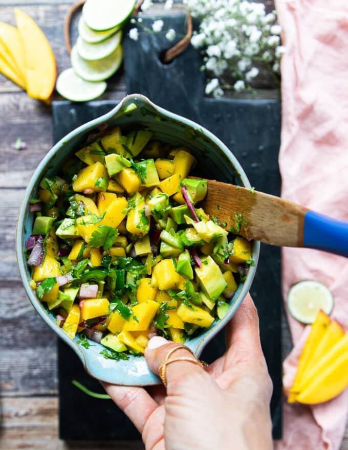 A hand holding a bowl of avocado salsa being prepared while the salmon is cooking