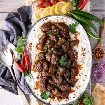 A large plate of lamb stew served over eggplant and yogurt to make Ali Nazik recipe, surrounded by red chilies, green chilies, lemon wedges, turkish pepper flakes, bread and tomatoes