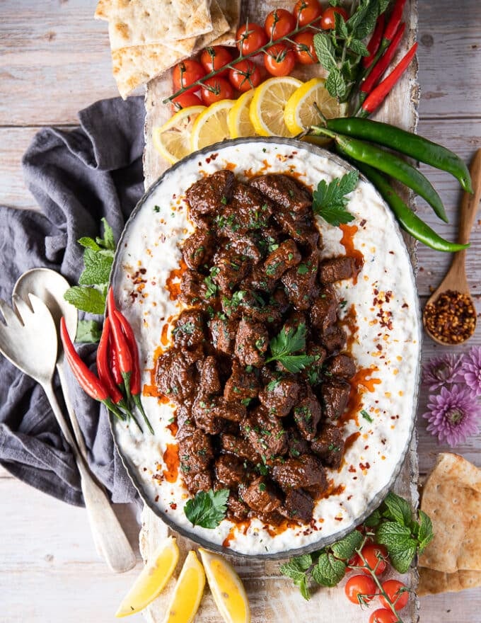 A large plate of lamb stew served over eggplant and yogurt to make Ali Nazik recipe, surrounded by red chilies, green chilies, lemon wedges, turkish pepper flakes, bread and tomatoes