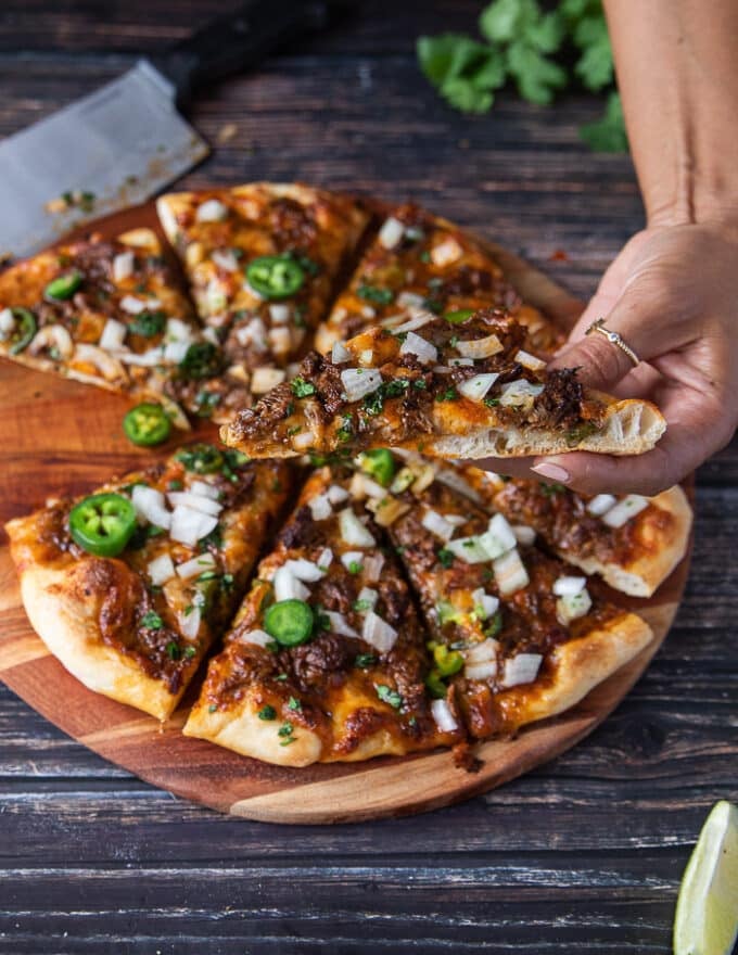 A hand picking up a slice of birria pizza to show the airy crisp crust and toppings