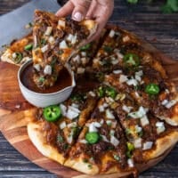 A hand dipping a slice of birria pizza into the birrita stew sauce