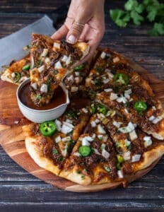 A hand dipping a slice of birria pizza into the birrita stew sauce