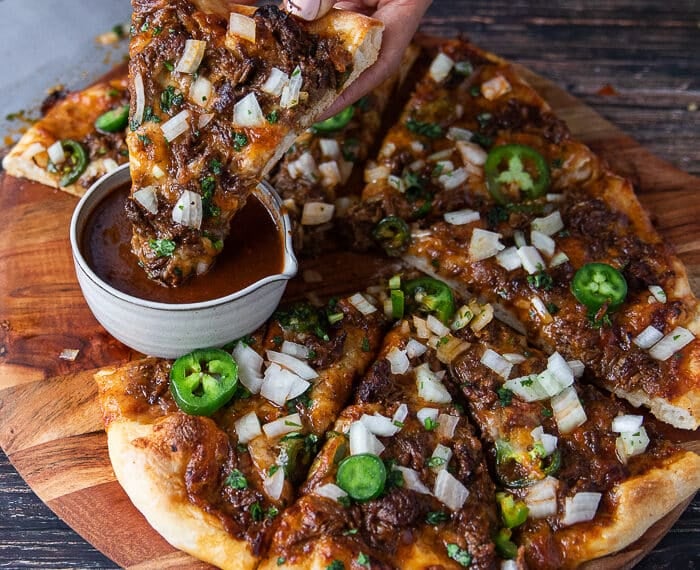 A hand dipping a slice of birria pizza into the birrita stew sauce