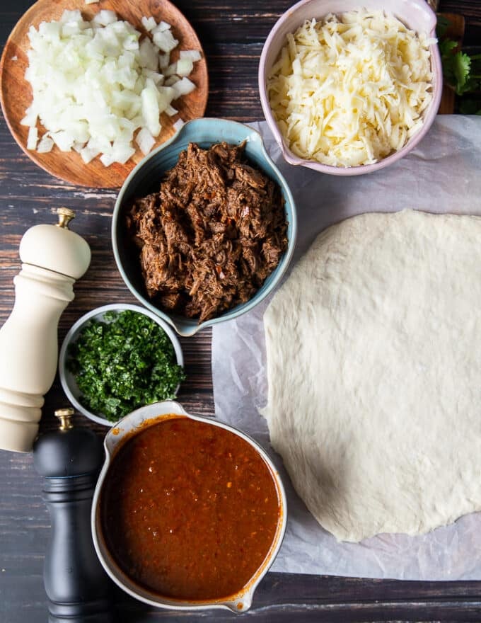 Zutaten für die Herstellung von Birria-Pizza, einschließlich: Pizzateig, Birria-Fleisch, Birria-Eintopf, Koriander, Jalapeños, Käse, Zwiebeln 