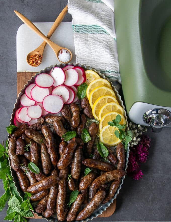 A plate of lamb sausage all cooked and plated with lemon slices, some radishes, fresh mint and sprinkled with chilli flakes