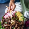 A hand holding a sausage link in a fork over a plate of lamb sausage