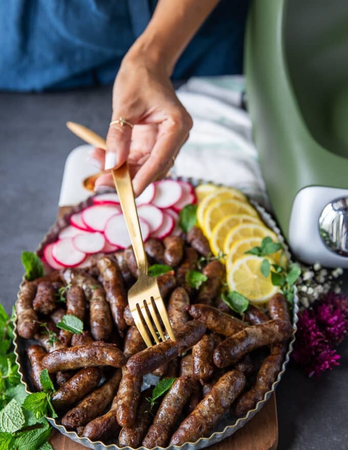 A hand holding a sausage link in a fork over a plate of lamb sausage