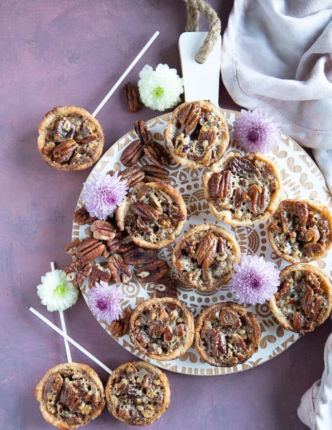 A fill plate of mini pecan pies, some with lollipop stick and some not. Surrounded by a tea towel and placed on a white board