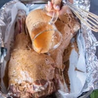 A hand pouring the wet marinade blend over the spice rubbed leg of lamb in the roasting pan