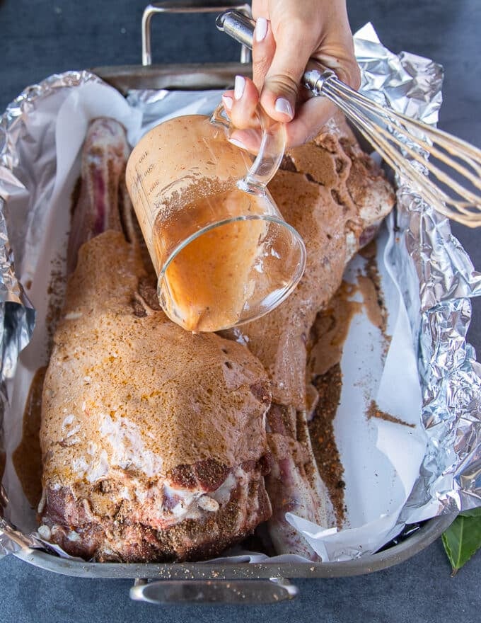 A hand pouring the wet marinade blend over the spice rubbed leg of lamb in the roasting pan