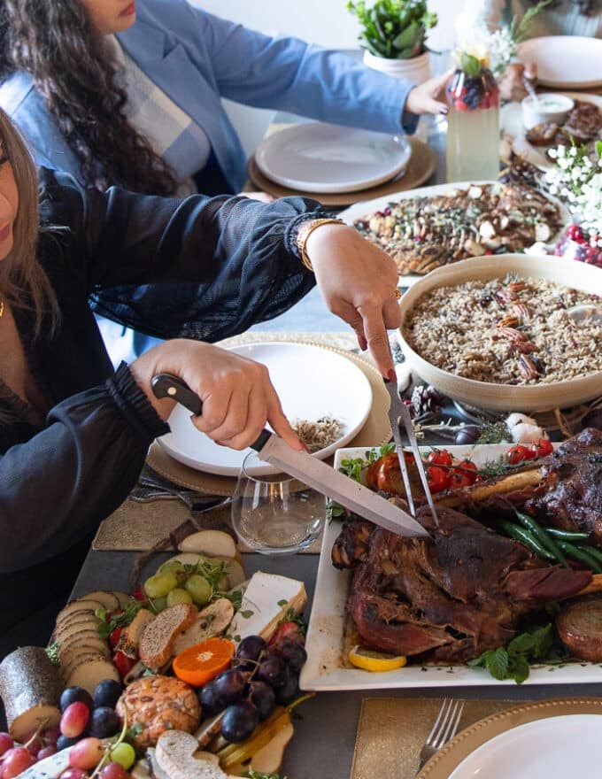 A whole dinner setup showing the leg of lamb served on a dining table along with rice, salad, cheese platter, roast potatoes and a hand slicing and carving the leg of lamb recipe