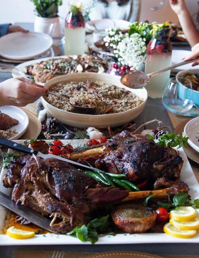 A table set up with leg of lamb recipe and hands serving up the lamb, the rice, the salad and enjoying a huge dinner
