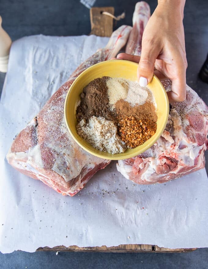 A hand holding a dry spice mix over fresh leg of lamb