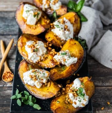 A plate of roasted acorn squash on a board topped with burrata cheese, toasted pine nuts and fresh herbs