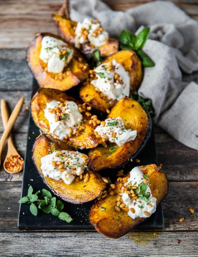 A plate of roasted acorn squash on a board topped with burrata cheese, toasted pine nuts and fresh herbs