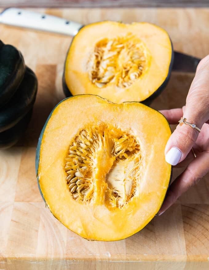 A hand holding a cut up piece of acorn squash showing the inside flesh and seeds before scooping