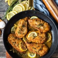 A skillet with chicken scallopini cooked up in it with lemon slices, capers and golden brown seared chicken breast