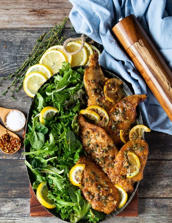 A plate of chicken scallopini on a wooden board served with greens and lemon slices surrounded by a black pepper mill