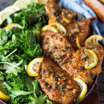 close up of chicken scallopini served on a plate with lemon slices, capers and some fresh parsley on top
