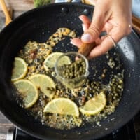 making the sauce for chicken scallopini: the chicken is removed from the skillet and more butter is added to the skillet along with lemon slices and a hand adding in capers