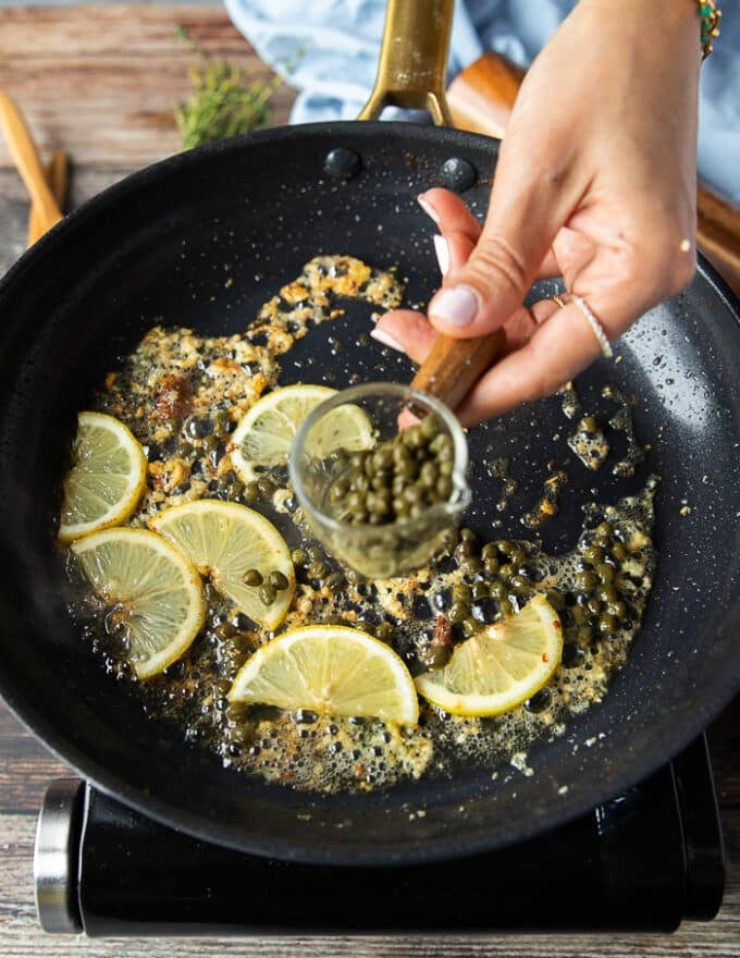 making the sauce for chicken scallopini: the chicken is removed from the skillet and more butter is added to the skillet along with lemon slices and a hand adding in capers