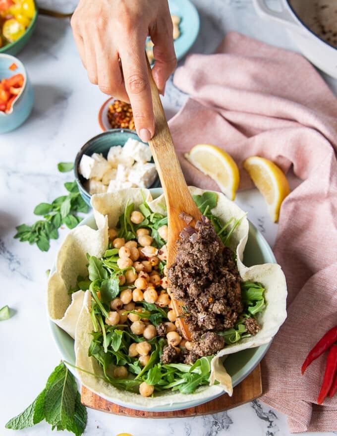A hand holding a wooden spoon and spooning the cooked lamb over the pita bowl with chickpeas and arugula