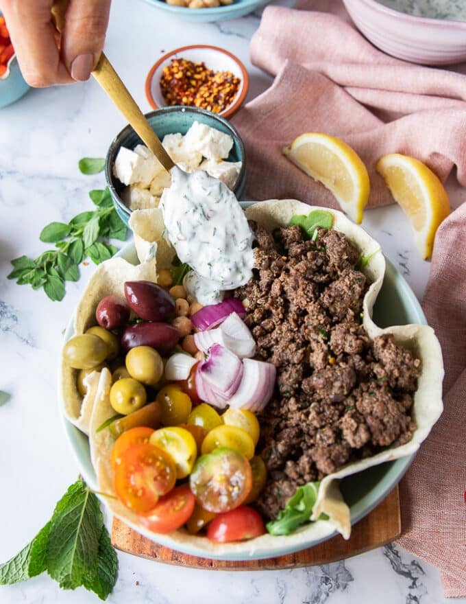 A hand dolloping the yogurt sauce on one side of the pita bowls which have been loaded with some tomatoes, onions, olives and cucumbers