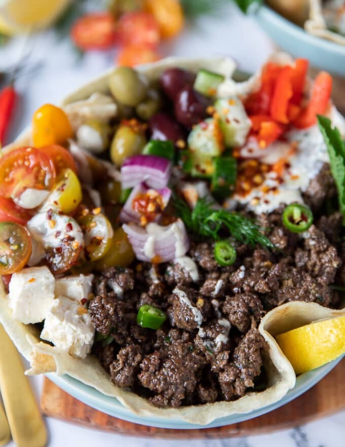 close up of one pita bowl showing the pita bread at the bottom and the cooked lamb, tomatoes, feta, yogurt, chickpeas, arugula, tomatoes, cucumbers, onions and chillies over the top