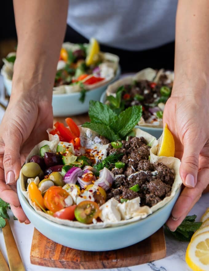 A hand holding one final assembled pita bowl showing the pita bread, the many toppings and sauces and a lemon wedge on top
