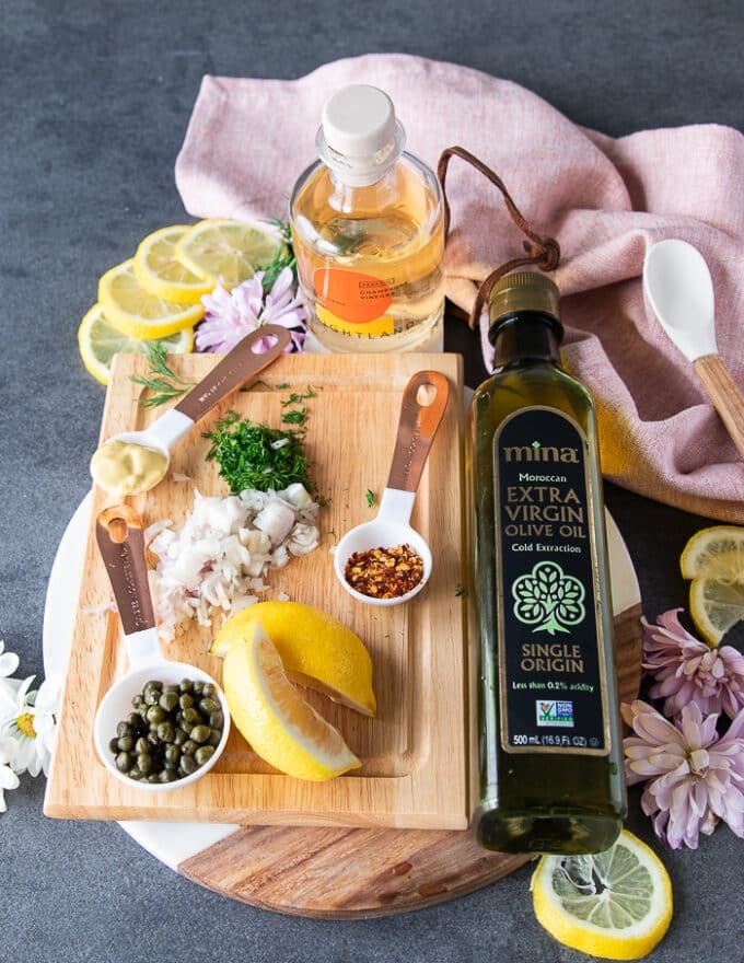 Ingredients for salmon tartare on a wooden board including the fresh salmon, a bowl of capers, a spoon of mustard, some chopped shallots, lemon wedges, fresh chopped dill, a bottle of olive oil, chili fakes and a bottle of vinegar