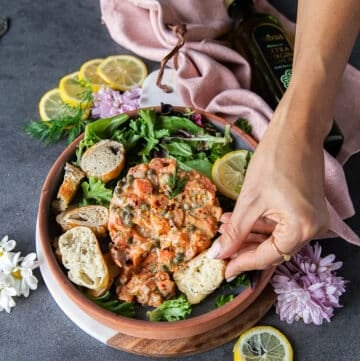 A hand holding a piece of bread and trying to poke through salmon tartare on a plate surrounded by greens and lemon slices