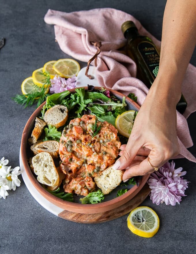 A hand holding a piece of bread and trying to poke through salmon tartare on a plate surrounded by greens and lemon slices