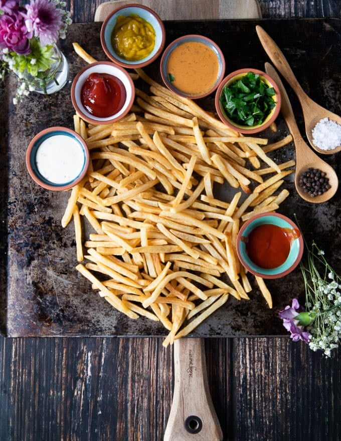 A large wooden board with the final air fryer frozen fries on a sheet surrounded by many bowls of dipping sauces, some salt and pepper and fresh herbs optional