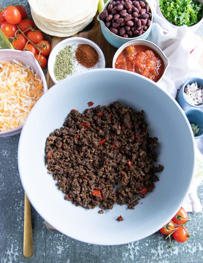 ingredients on a bowl to make taquitos include: a bowl of cooked beef mixture, a bowl of black beans, a bowl of shredded cheese, a bowl of tomatoes diced, a bowl of cilantro minced, a bowl of garlic minced, shallot minced and homemade salsa
