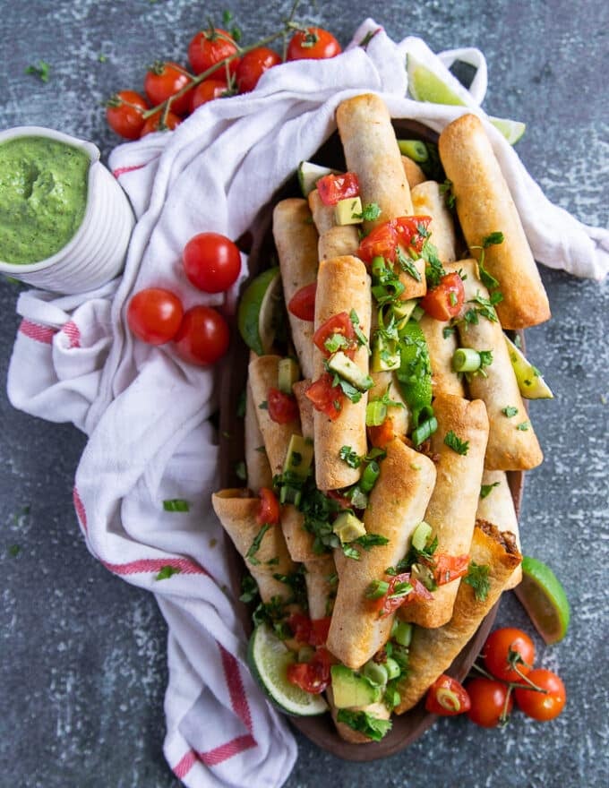 A large serving plate of taquitos surrppunded by a bowl of poblano sauce, some fresh tomato salsa, some lime wedges and a tea towel