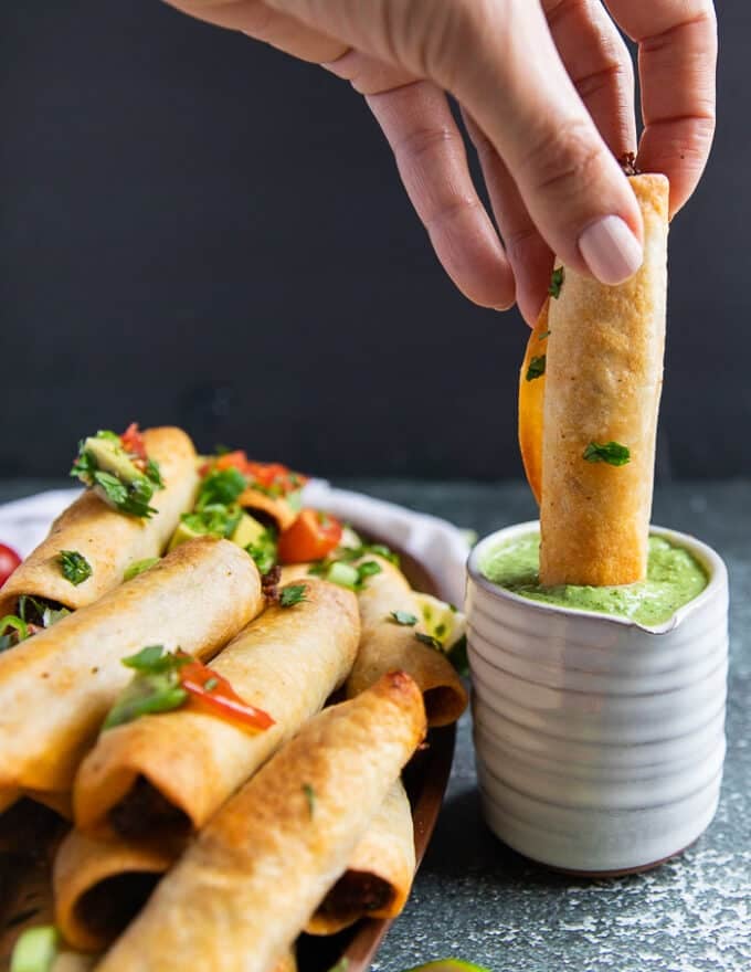 A hand dipping one taquito into a bowl of poblano sauce 