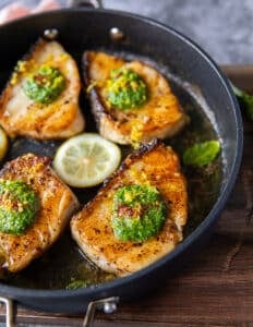 Chilean Sea bass cooked in a skillet with a gorgeous golden crust surrounded with lemon slices and topped with a dollop of basil pesto