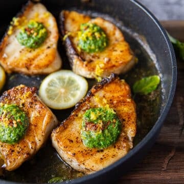 Chilean Sea bass cooked in a skillet with a gorgeous golden crust surrounded with lemon slices and topped with a dollop of basil pesto