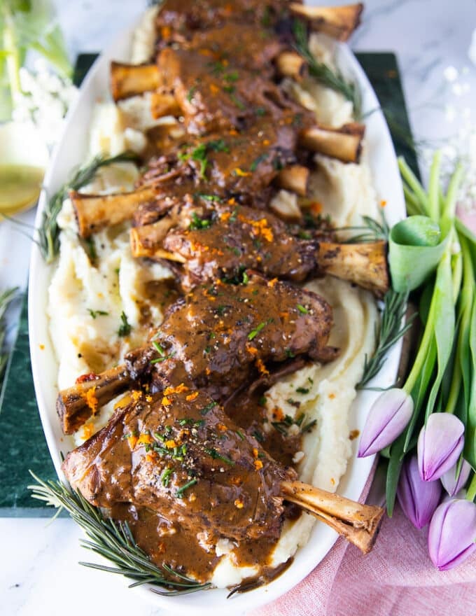 Some braised lamb shanks garnished with orange zest and fresh rosemary over a plate of mashed potatoes 
