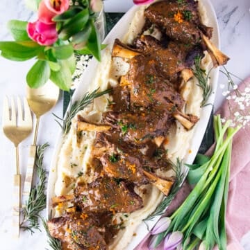 A large plate of braised lamb shanks with gravy over mashed potatoes surrounded by fresh tulips and a serving spoon