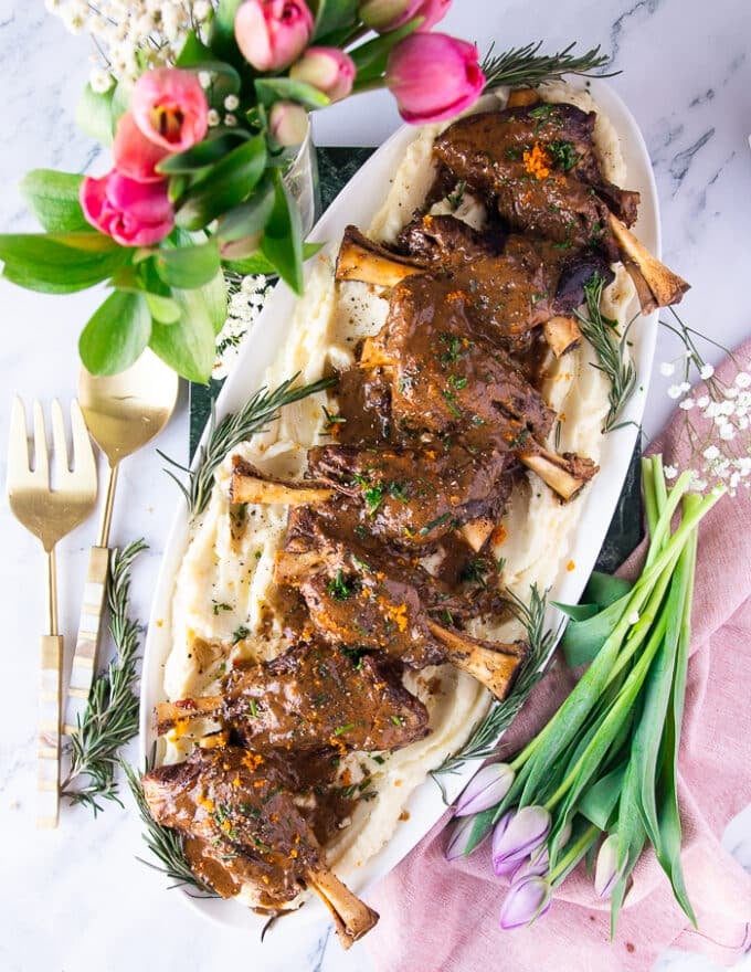 A large plate of braised lamb shanks with gravy over mashed potatoes surrounded by fresh tulips and a serving spoon