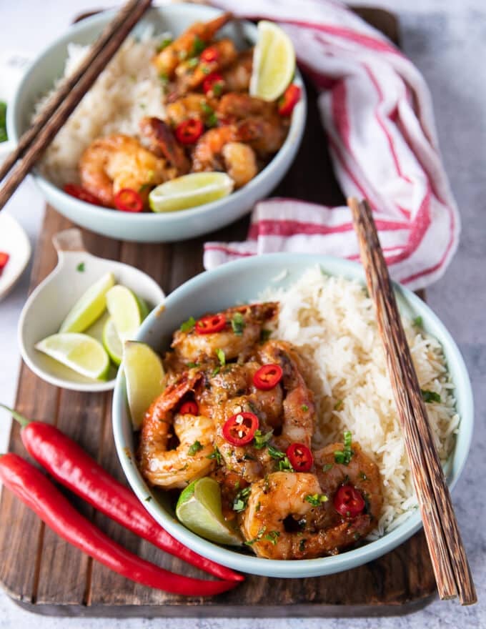 two plates of chili lime shrimp served over rice on a wooden board with chop sticks and lime wedges