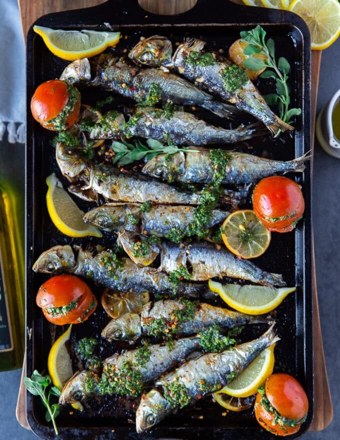 Baked sardines on a baking sheet with lemon slices, a drizzle of chermoula sauce and some stuffed tomatoes on the side