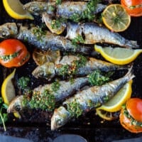 close up of a cooked sardine on a sheet with lots of herb sauce chermoula and lemon wedges