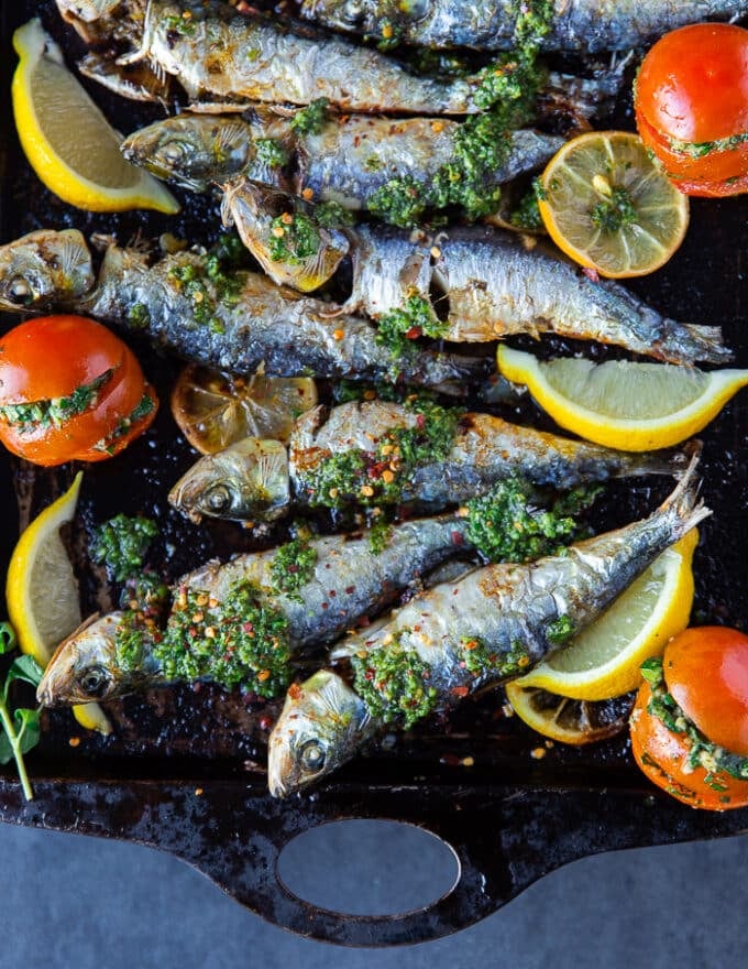 close up of a cooked sardine on a sheet with lots of herb sauce chermoula and lemon wedges