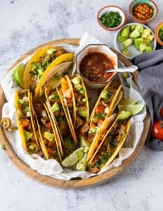 A large plate of ground chicken tacos loaded with avocados, tomatoes, onions, alongside a bowl of homemade salsa and lime wedges