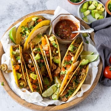 A large plate of ground chicken tacos loaded with avocados, tomatoes, onions, alongside a bowl of homemade salsa and lime wedges