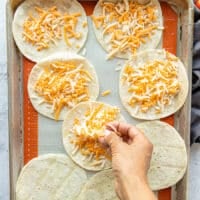 A hand sprinkling a generous amount of cheese on the soft shell tortillas on a baking sheet ready to melt in the oven