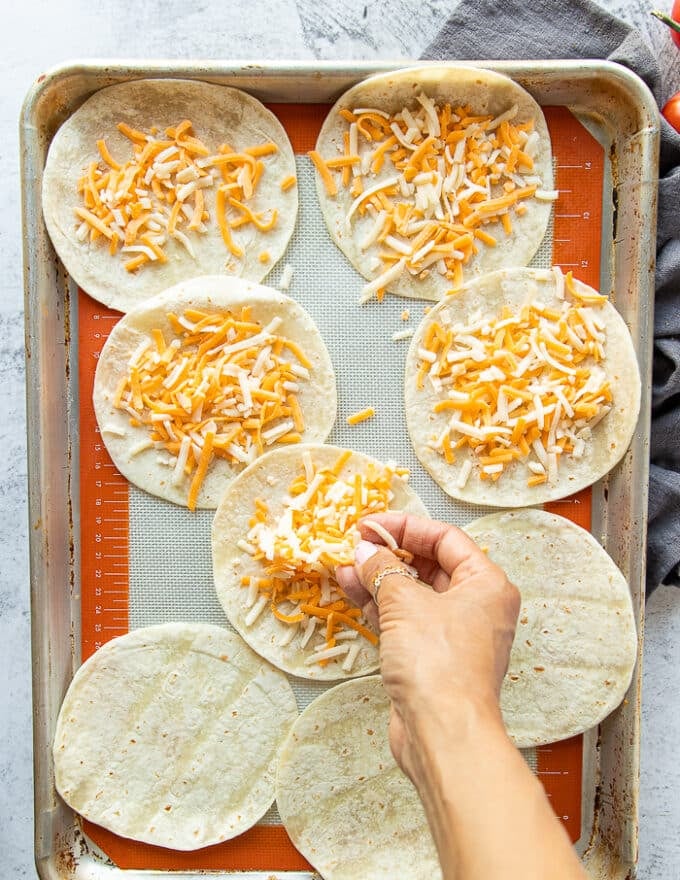 A hand sprinkling a generous amount of cheese on the soft shell tortillas on a baking sheet ready to melt in the oven