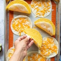 Once the cheese melted on the same baking sheet, a hand placing a hard shell tortilla and sticking it to the soft cheesy shell by pressing it down.